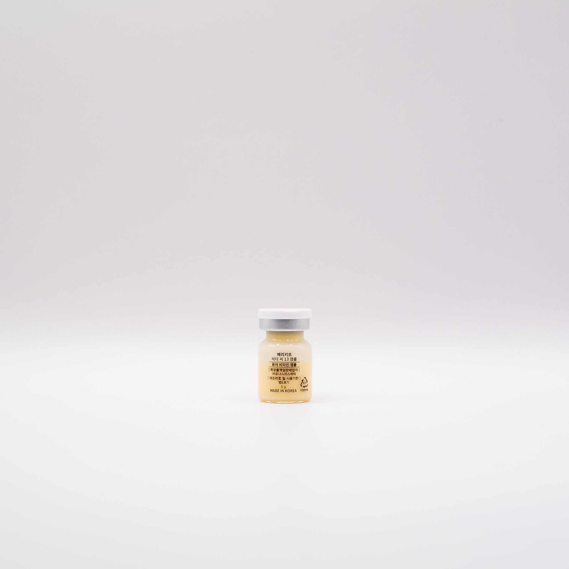 Single small clear glass ampoule bottle with white cap containing MERIKIT Vita-C 13 vitamin C serum against a plain white background